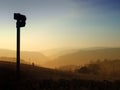 Direction sign in silhouette pointing down a glowing evening sunset valley with golden tree covered hills at twilight