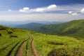 Dirt car track on green grassy hill leading to woody mountains ridge on bright blue sky copy space background. Tourism and Royalty Free Stock Photo