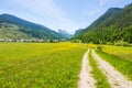 Dirt country road crossing flowery meadows, mountains and forest in scenic alpine landscape and moody sky. Summer adventure and Royalty Free Stock Photo