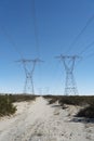 A dirt road following between the path to two high tension power towers and lines stretching across desert. Royalty Free Stock Photo