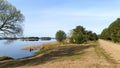 A dirt road runs along the grassy shore of the lake with pines and willows growing there. The calm water reflects the blue sky and Royalty Free Stock Photo