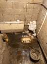 Dirty toilet room with pile of used toilet paper and a bucket, uncoated concrete flooring and cinder brick walls.