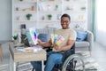 Disabled black guy in wheelchair working as graphic designer online, holding color swatches palette, using laptop indoor