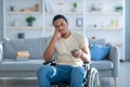 Disabled black man in wheelchair using remote control, watching dull movie on TV, cannot find interesting program Royalty Free Stock Photo