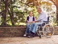 Disabled grandfather in wheelchair in park spending time together with his granddaughter reading book. Royalty Free Stock Photo