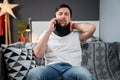 Disabled man with neck brace talking on phone with doctor, experiencing pain while sitting on couch in the living room Royalty Free Stock Photo