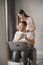 Young man in wheelchair using laptop and girlfriend hugging him at home