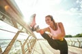 Disabled positive woman in sportswear and headphones stretching prosthetic leg while standing on the bridge. Listening