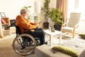 Disabled senior man sitting on wheelchair in cozy living room on a daytime and using mobile phone for news reading and Royalty Free Stock Photo