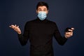 Disapointment young man in black sweater, with sterile face mask spreading hand with mobile phone, isolated on Pacific Blue