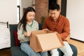 Disappointed asian married spouses unpacking cardboard box, received damaged item or unwanted stuff, sitting on sofa