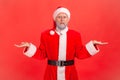 Disappointed confused gray bearded elderly man in eye glasses and santa claus costume throws up hands, cant understand how to