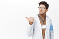 Disappointed doctor in robe with name tag, pointing left and looking displeased, face of dislike, standing over white