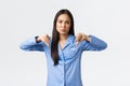 Disappointed frowning asian woman in blue pajamas express dislike, showing thumbs-down skeptical, judging bad product or