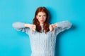 Disappointed redhead girl in sweater showing thumbs-down, judging bad product, disagree and dislike promo, standing over