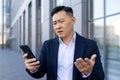 A disappointed young Asian businessman spreads his hands while looking at the mobile phone screen, standing outside an Royalty Free Stock Photo