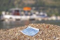 A discarded medical face mask contaminates a beautiful sandy pebble beach, with fishing boats in the background. Budva, Montenegro