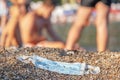 Discarded used face mask lies on a sandy pebble beach, beachgoers are relaxing by the sea in the background. Budva, Montenegro Royalty Free Stock Photo