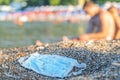 Discarded used face mask lies on a sandy pebble beach, beachgoers are relaxing by the sea in the background. Budva, Montenegro Royalty Free Stock Photo