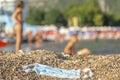 Discarded used face mask lies on a sandy pebble beach, beachgoers are relaxing by the sea in the background. Budva, Montenegro Royalty Free Stock Photo