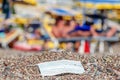 Discarded used facial mask lies on a sandy pebble beach, in the background beachgoers are relaxing by sun loungers and beach Royalty Free Stock Photo