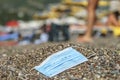 Discarded used facial mask lies on a sandy pebble beach, in the background beachgoers are relaxing by sun loungers and beach Royalty Free Stock Photo