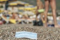 Discarded used facial mask lies on a sandy pebble beach, in the background beachgoers are relaxing by sun loungers and beach Royalty Free Stock Photo
