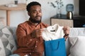 Discontented African American Male Buyer Unpacking Shopper Bag At Home