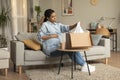 Discontented young pregnant black woman unpacking box and holding delivered clothes, sitting on sofa at home, free space Royalty Free Stock Photo