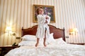 Discover happiness here. Cheerful boy in white bathrobe is having fun, jumping on the white bed in a hotel room. The kid