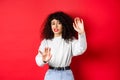 Disgusted young woman begging to stop, raising hands defensive, say no, standing on red background