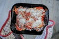 Dish of aubergine parmigiana ready to put in the oven. Black dish being held by oven gloves