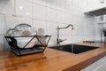 Dish rack holds many dishes and cups against wooden countertop, white wall tiles, sink and faucet. Budget and lightweight