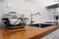 Dish rack holds many dishes and cups against wooden countertop, white wall tiles, sink and faucet. Budget and lightweight