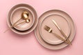 Dishes and utensils for serving and eating meals. Beige round rimmed plates and golden cutlery on a pink background, top view.