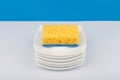 Dishwashing concept, high angle view of cleaning sponge on pile of clean saucers on white table with blue background