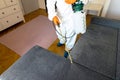 Person in protective suit with decontamination sprayer bottle disinfecting household and furniture