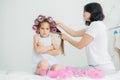 Displeased child keeps hands crossed, looks with sullen expression, being discontent as her mother winds curlers on her hair, does