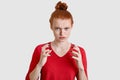 Displeased foxy young woman has freckled skin, keeps hands in angry gesture, looks with anger, wears red clothes, isolated over