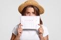 Displeased woman holds perids calendar, wears straw hat, has discontent expression, dressed in casual t shirt, has problems with w