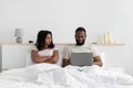 Displeased young black wife with crossed arms looks at busy husband, working on laptop, sitting on bed in white bedroom Royalty Free Stock Photo