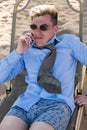 Disgruntled young businessman downshifter talks on the phone lying on a sun lounger on the beach in a shirt, tie and