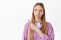 Displeased young woman smirk and frown, looks and points finger left at something disappointing, feeling regret or Royalty Free Stock Photo