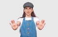 Displeased ypung woman wearing blue shirt, gray cap and round transparent eyewear, posing against gray studio wall, keeping hands