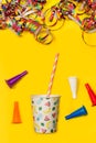 A disposable cup with a striped straw, serpentines and toy cornets on a yellow background