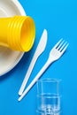 Disposable plastic tableware on blue background. White plate forks, knives, yellow and transparent plastic glasses Royalty Free Stock Photo