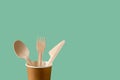 Disposable wooden spoon, fork and knife in a brown paper cup against green background. Biodegradable tableware for takeaways, Royalty Free Stock Photo