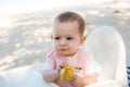 Dissatisfied baby girl sits and eats banana on a white kids chair