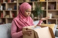 Dissatisfied black muslim woman unpacking cardboard box, receiving damaged item, sitting on sofa at home, copy space Royalty Free Stock Photo