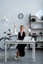 Dissatisfied businesswoman in formal wear sitting at desk, gesturing with hands and throwing documents in air